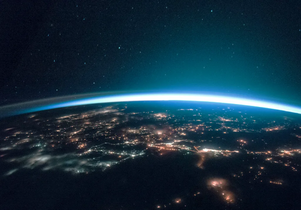 View of Earth from space at night, showing city lights and the thin glowing atmosphere against a dark sky.